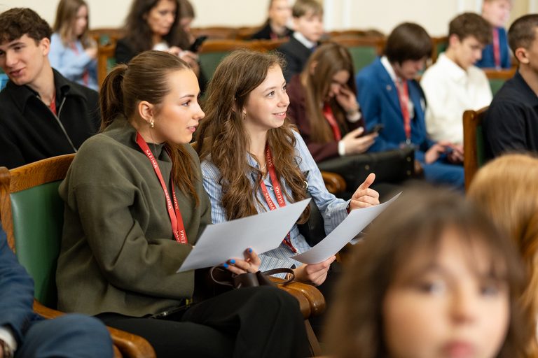 Divas jaunietes ar sarkanām kakla lentēm ar uzrakstu "SAEIMA" sēž zālē starp citiem pasākuma dalībniekiem, tur rokās papīra lapas un vērīgi klausās.