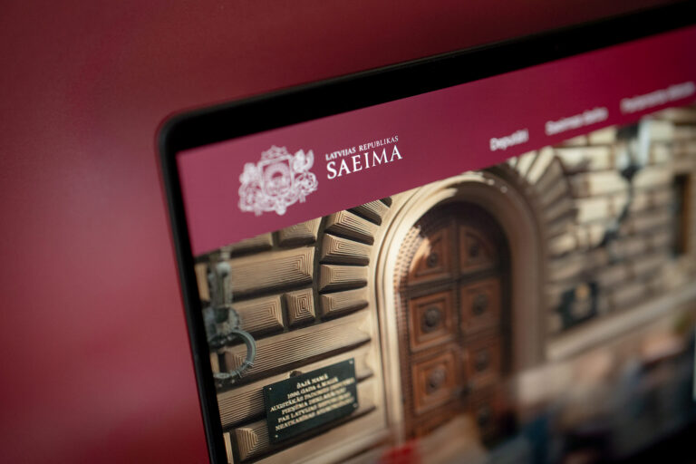 Klēpjdatora ekrāns leņķī ar atvērtu tīmekļa vietni, kuras augšējā bordo krāsas joslā redzams Latvijas ģerbonis un teksts "LATVIJAS REPUBLIKAS SAEIMA", bet fonā – Saeimas nama fasāde ar ieeju un piemiņas plāksni.