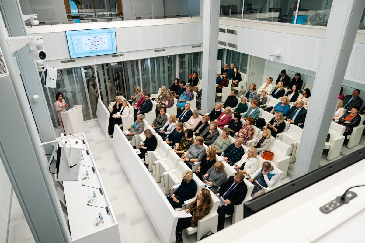 Skats no augšas uz modernu, baltu konferenču zāli, kurā pie tribīnes uzstājas referente, bet rindās izkārtotos baltos sēdekļos klausās plaša auditorija. Virs zāles pie sienas novietots ekrāns ar prezentācijas shēmu.