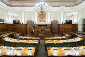 Latvijas Republikas Saeimas Sēžu zāle ar puslokā izvietotām deputātu darba vietām, prezidija tribīni centrā, Latvijas un Eiropas Savienības karogiem, greznu lustru un pie kreisās sienas redzamu uzrakstu: LATVIJAS VALSTS SUVERĒNĀ VARA PIEDER LATVIJAS TAUTAI.