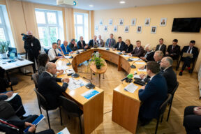 Deputāti lietišķā apģērbā sēž ap pakavveida koka galdu sapulču telpā ar dokumentiem un mikrofoniem priekšā, kamēr malā stāv videooperators ar kameru uz statīva.