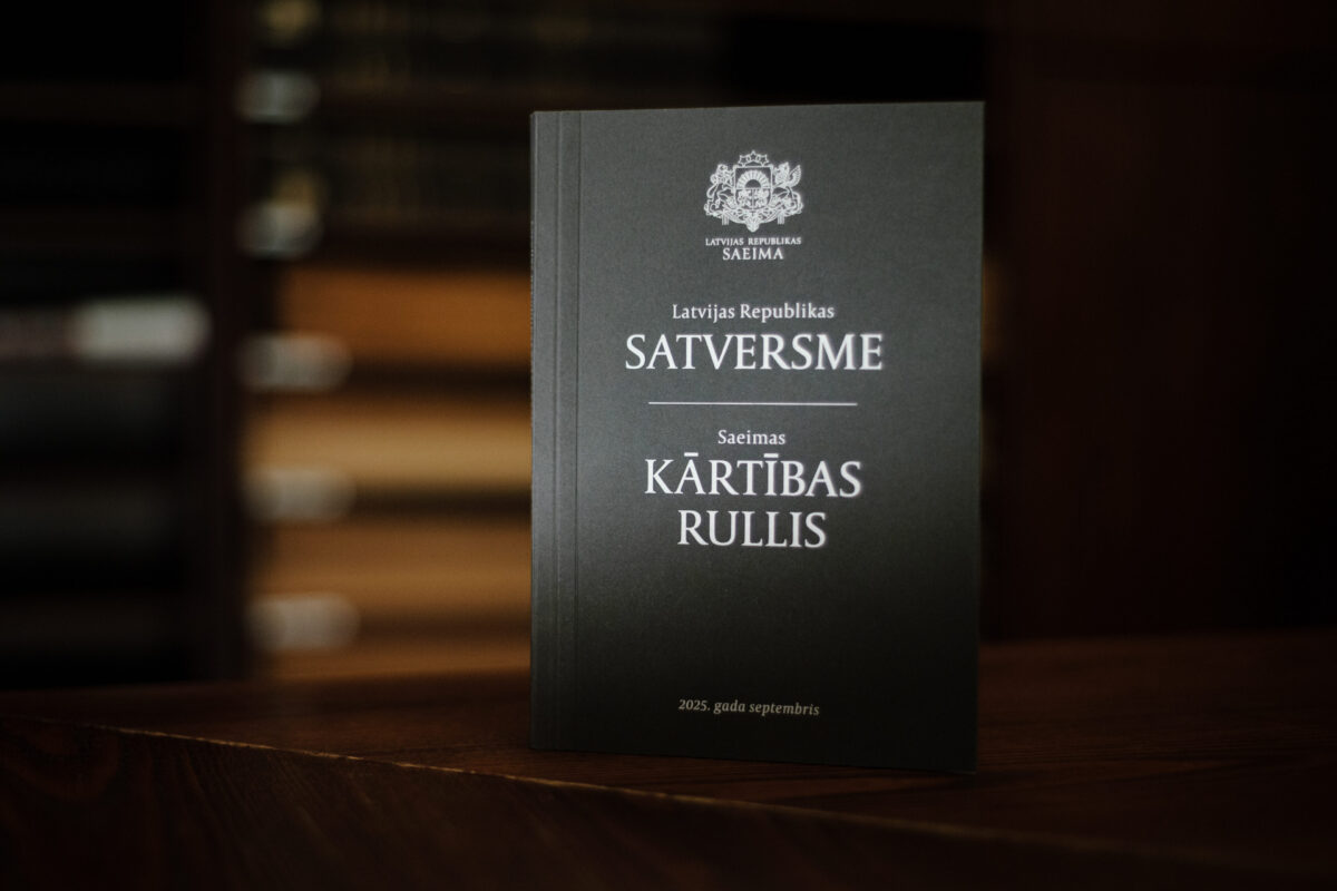 Uz koka virsmas novietota tumša grāmata ar Latvijas valsts ģerboni un tekstu: „LATVIJAS REPUBLIKAS SAEIMA”, „Latvijas Republikas SATVERSME”, „Saeimas KĀRTĪBAS RULLIS” un apakšā „2023. gada septembris”. Fonā redzams izplūdis grāmatplaukts.