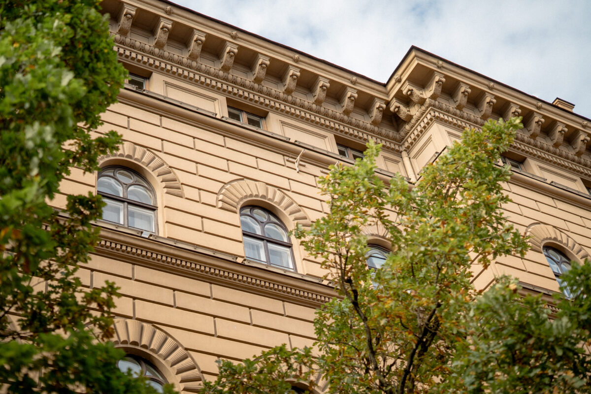 Skats no apakšas uz Saeimas ēkas smilškrāsas fasādi ar arkveida logiem un dekoratīvu karnīzi, ko daļēji ieskauj zaļu koku zari.