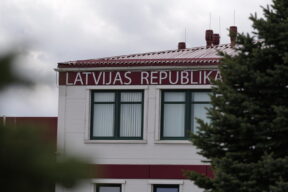 Ēkas fasādes augšdaļa ar uzrakstu “LATVIJAS REPUBLIKA”. Attēlā redzami vairāki logi, sarkanīgs metāla jumts un daļēji koku zari priekšplānā. Fonā apmākušās debesis.